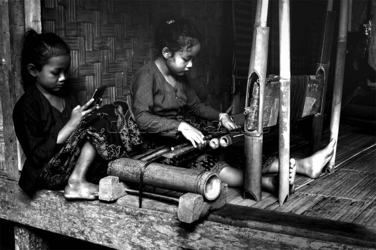 Baduy girl weaving traditional fabric while another child uses a smartphone in a wooden house, Banten, Indonesia