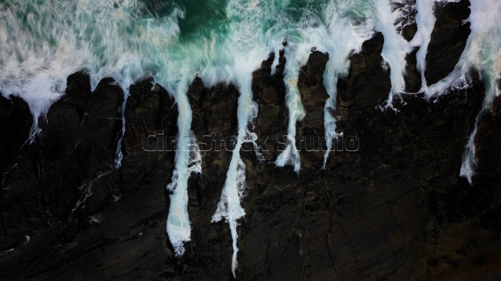 Top view long exposure ocean waves crash against the black rocks.