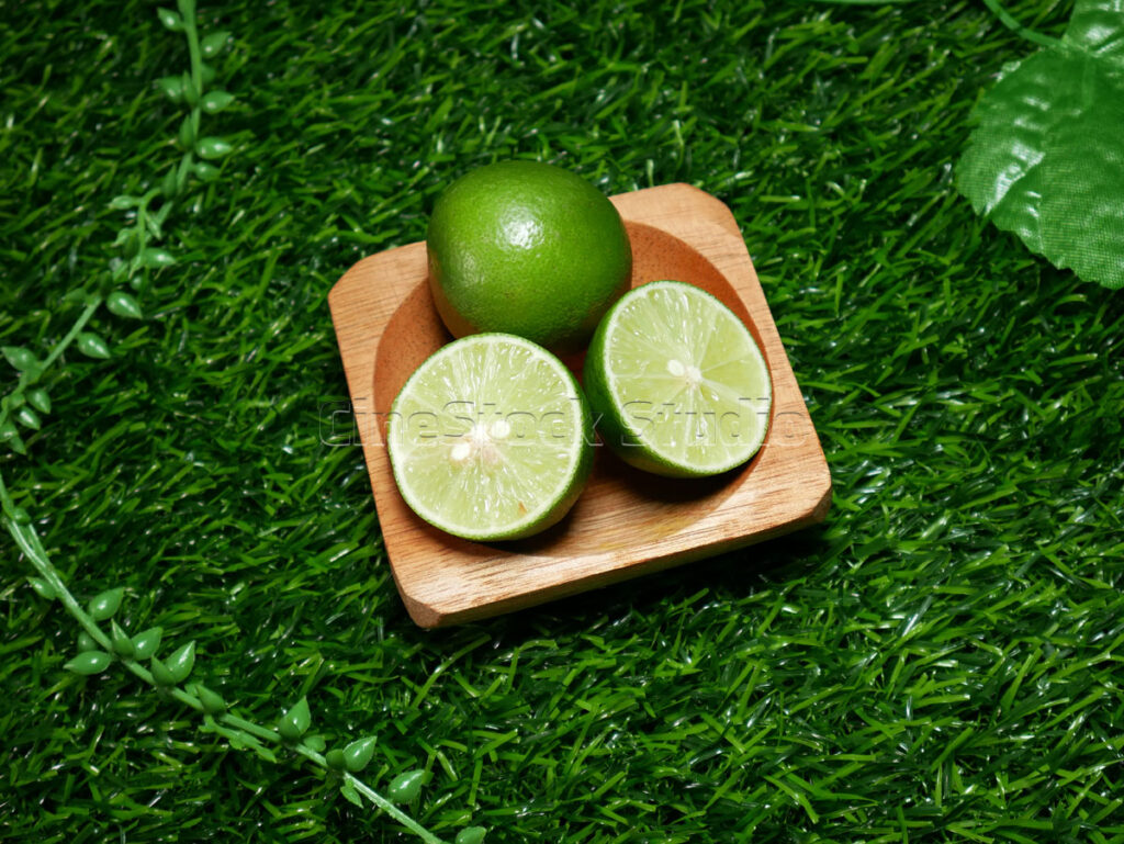 Fresh green lime slices and a whole fruit on a small wooden tray over a lush grass background.