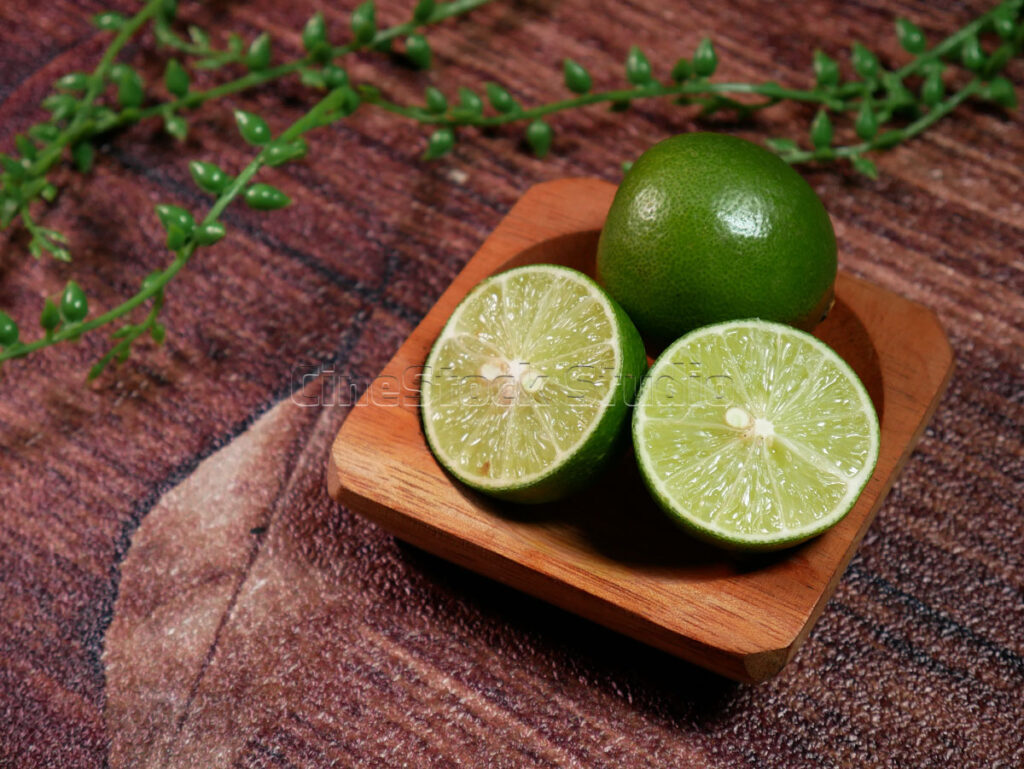 Organic lime fruits presented on a wooden plate against a dark rustic wood surface with decorative leaves.