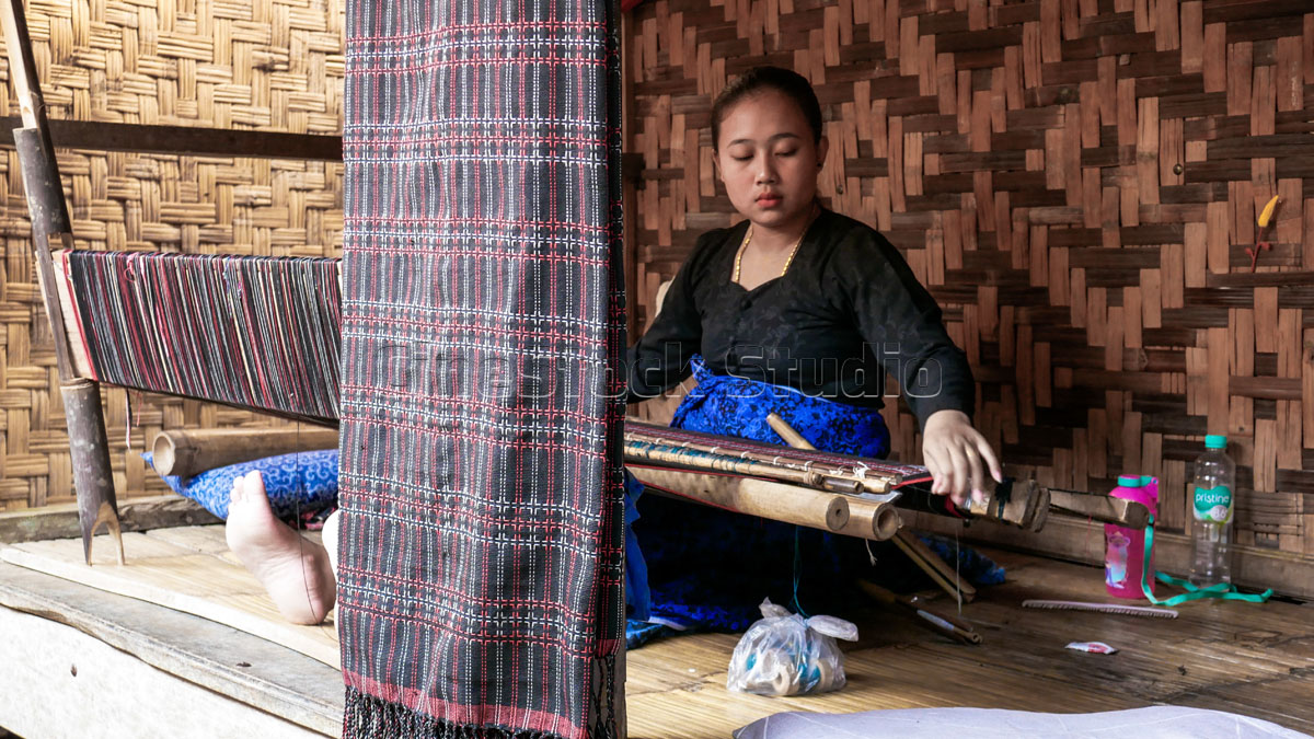 Indonesian Face A Baduy woman in traditional black and blue attire weaving on a wooden porch