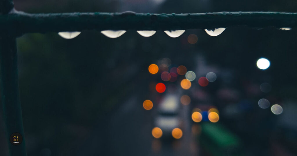 Featured image showing water droplets on an iron fence railing with a blurred bokeh background of city traffic lights after the rain.