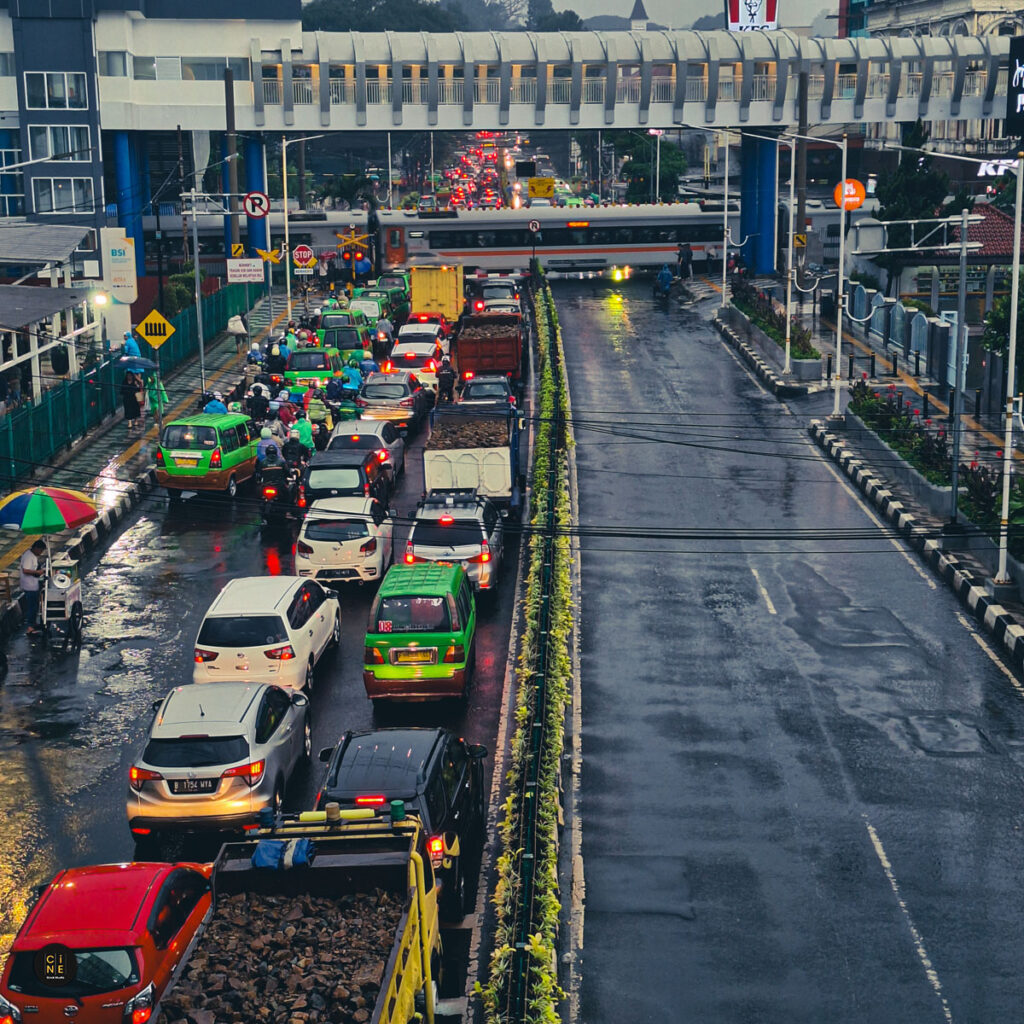 A high-angle editorial view of snaking vehicle traffic and crowded streets on damp asphalt near a pedestrian bridge.