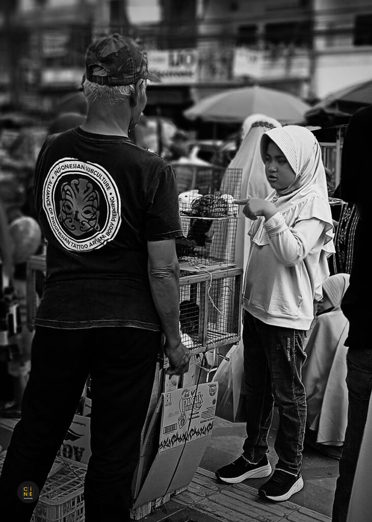 A candid black and white shot of a young girl standing in front of a wooden rabbit cage talking to the vendor.