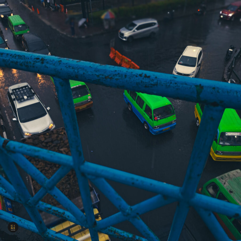A high-angle shot looking through blue iron pedestrian overpass railings at green angkot vans and cars on a wet road.