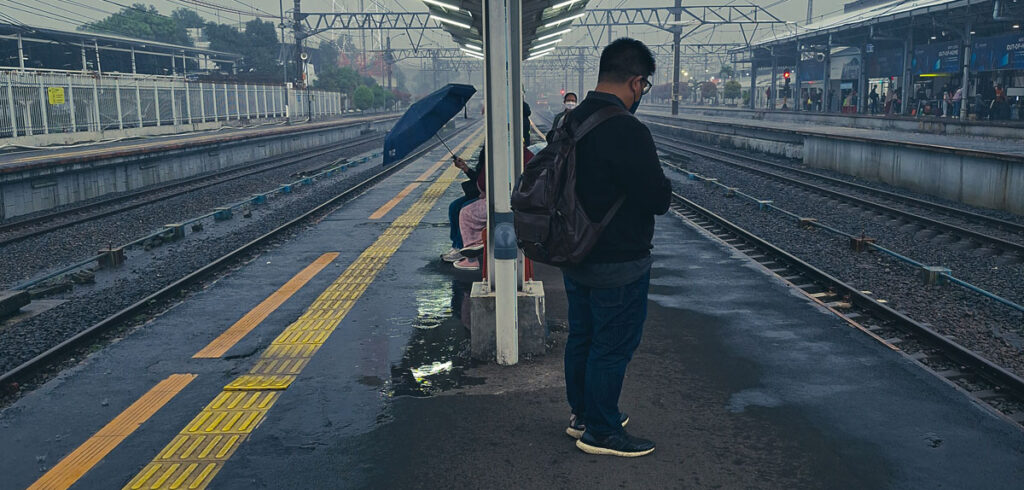 A monochrome-toned street photograph of a lone passenger standing on a wet platform at Bogor railway station.