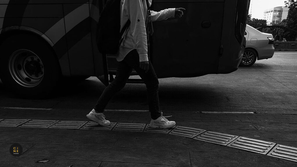 A cropped monochrome street shot of a person's legs in mid-stride, walking past the wheels of a large transit bus.