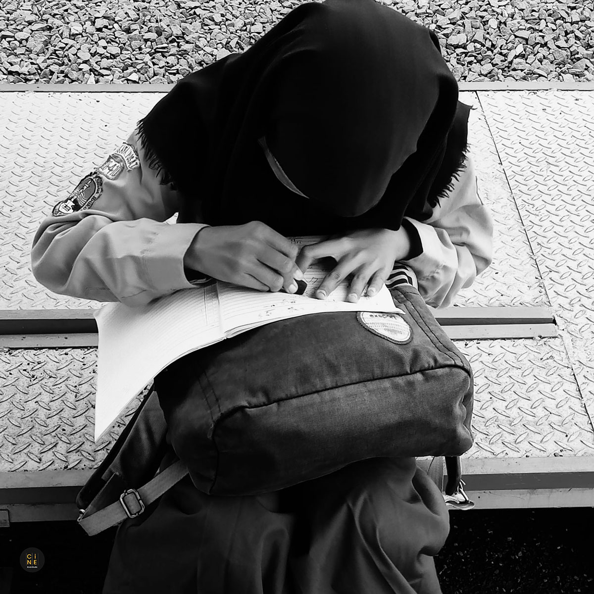 A close-up monochrome shot of a student deeply focused on writing her schoolwork using a backpack as a desk.