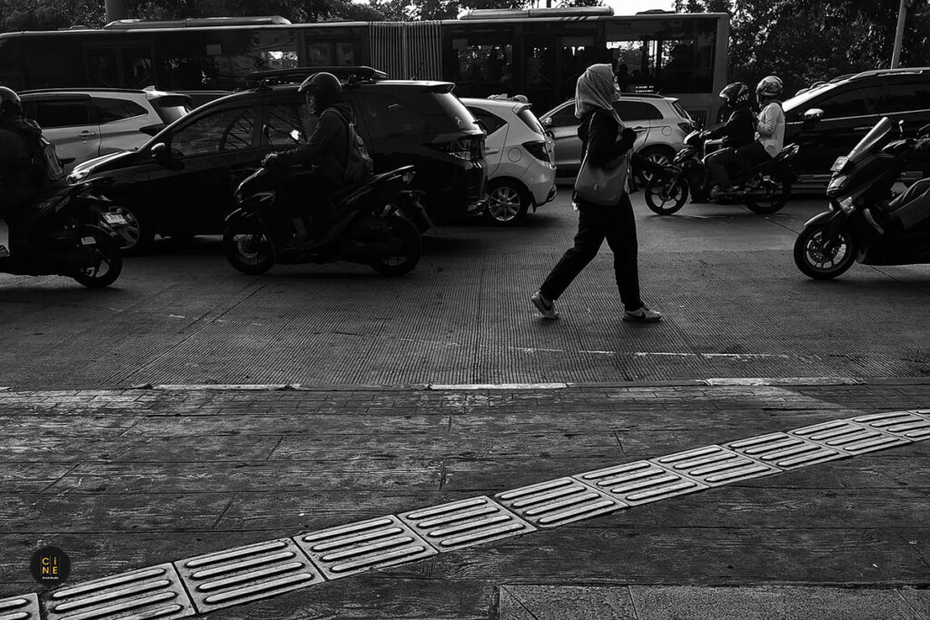 A black and white street photograph of heavy motorcycle and car traffic passing by a busy roadside sidewalk.