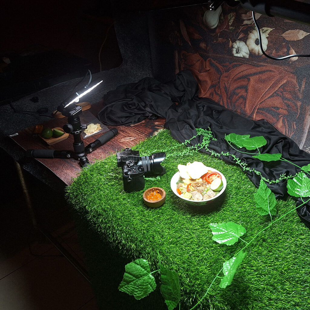 An overhead flat-lay view of a dark studio table where a Lumix G85 camera on a mini tripod shoots a bowl of food.