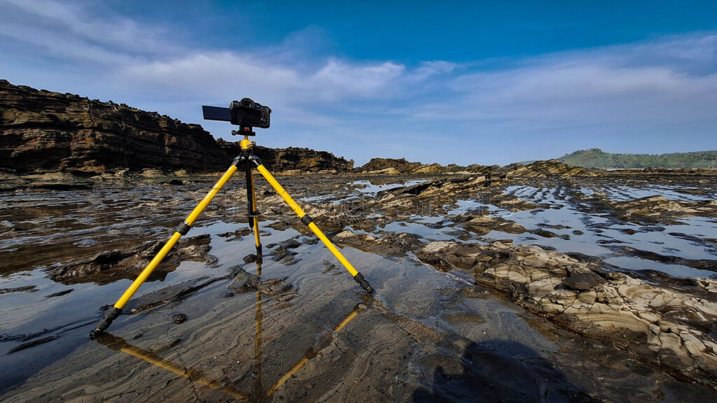 A professional mirrorless camera setup on a yellow tripod overlooking a wide rocky reef beach under a clear blue sky.