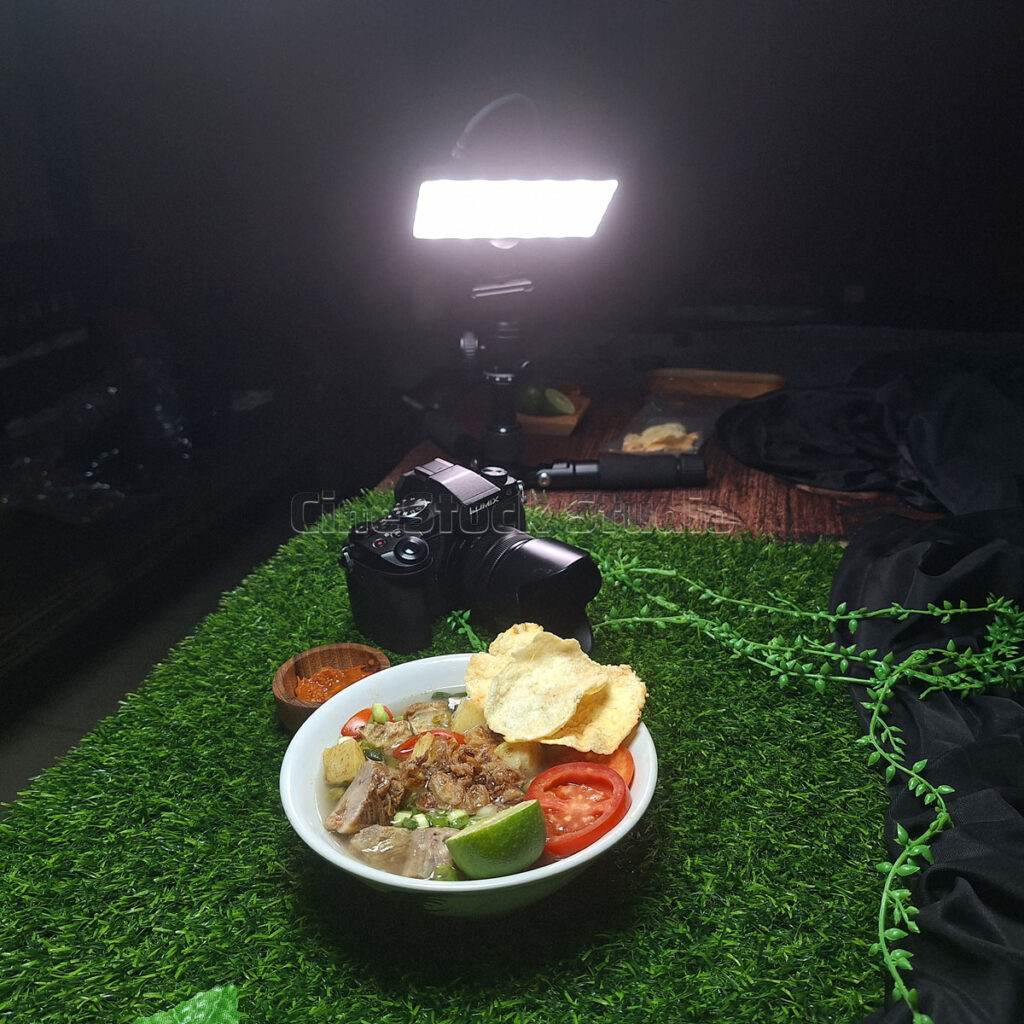 A dark studio environment showing an LED light panel illuminating a bowl of food from above, with a Lumix G85 nearby.