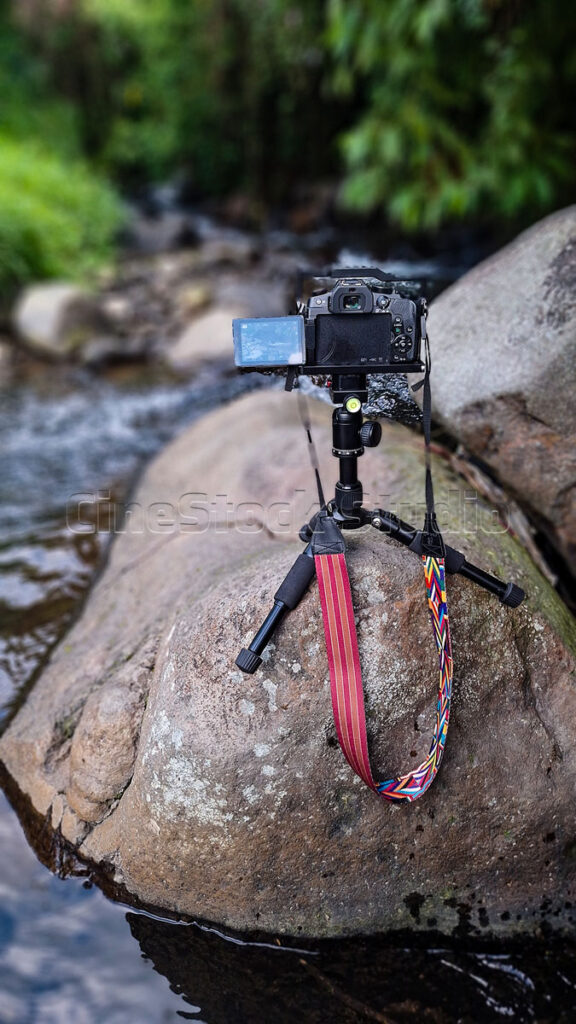 A vertical shot of the Lumix G85 camera on a tripod next to a large boulder, capturing a flowing river stream with a soft green background.