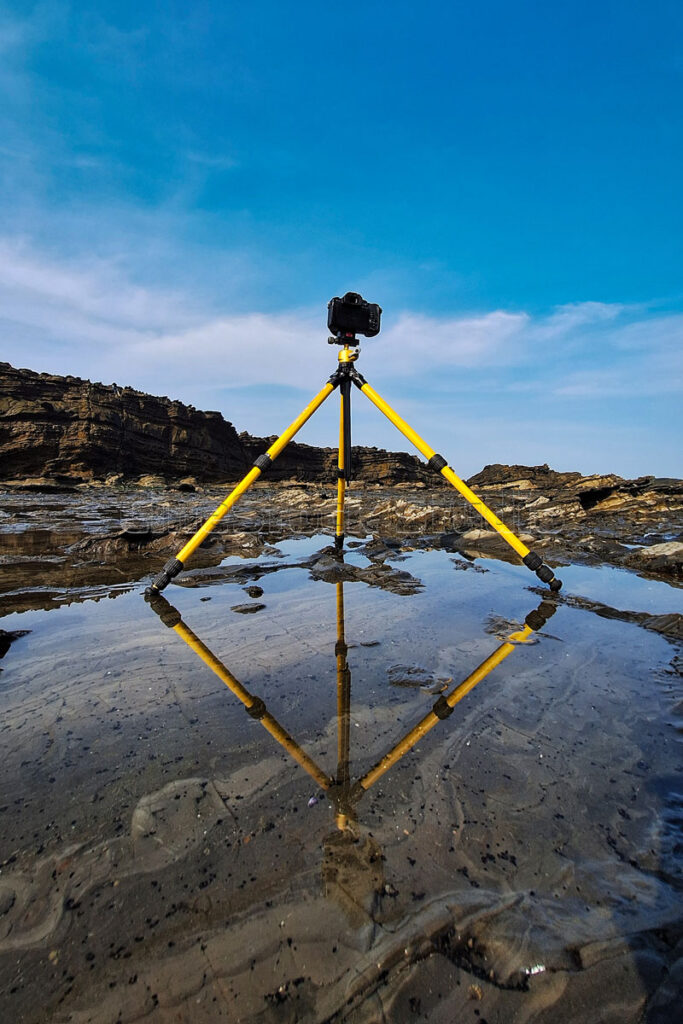 A vertical composition of a camera rig creating a perfect reflection on a still puddle of sea water along the rocky coast.