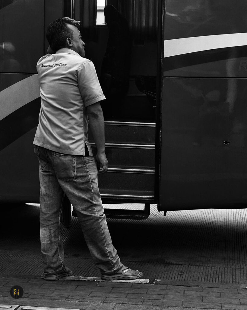 A vertical monochrome photograph of a man standing alone, looking toward the side of a large parked public bus.