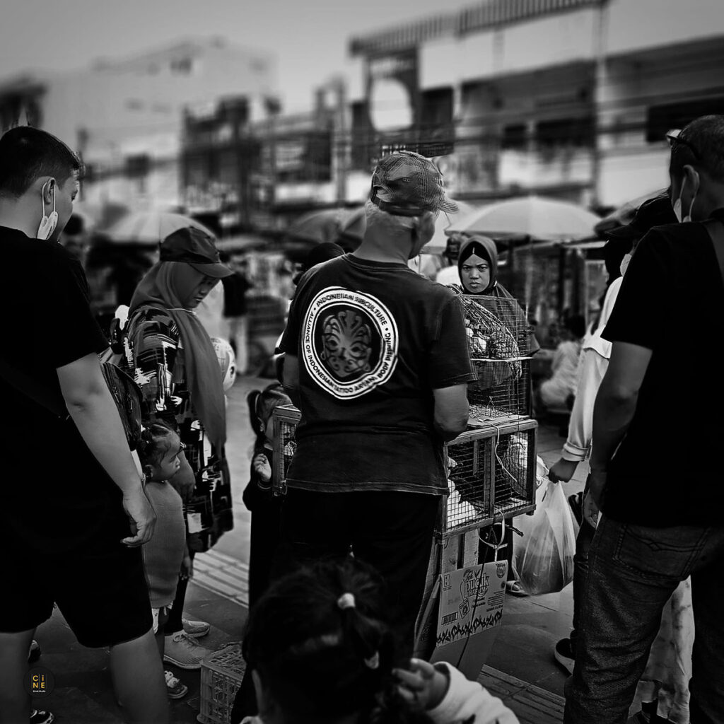 A black and white street photograph of a rabbit vendor from behind, surrounded by people in a crowded public square.