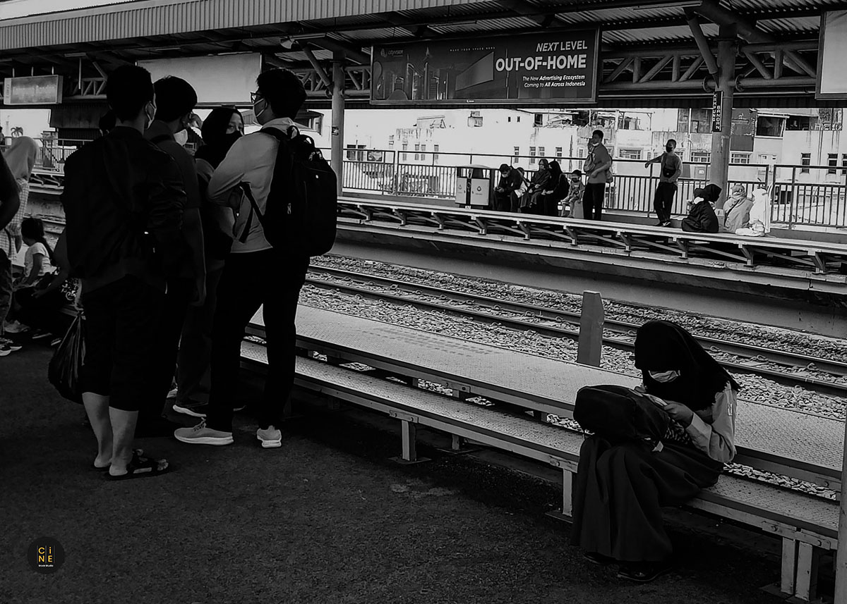 A group of passengers standing on the platform while a student sits alone studying in the background.