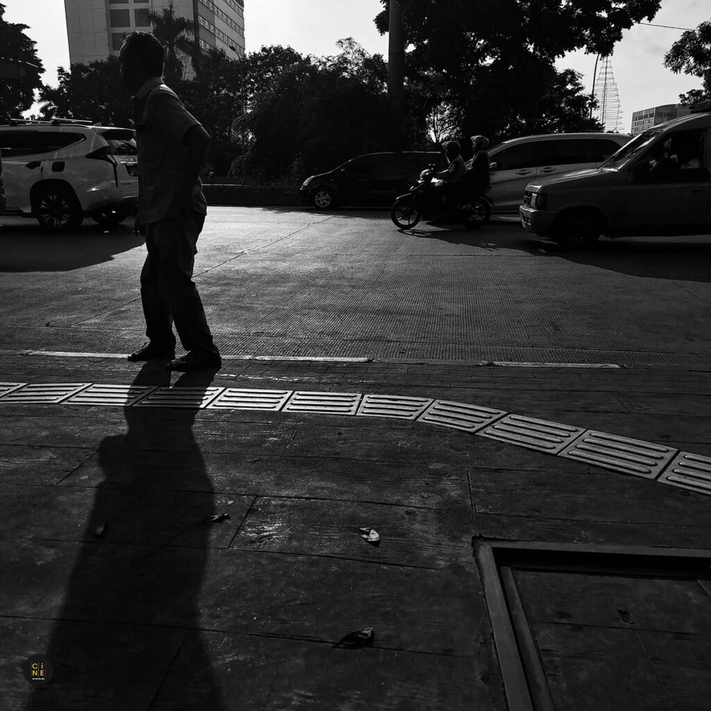 A high-contrast black and white shot of a person walking, casting a long dramatic shadow on the concrete ground.