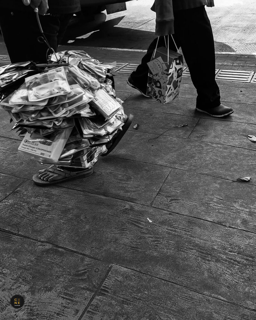 Close-up monochrome shot of a street vendor's feet walking while carrying a large bundle of goods in a plastic wrapper.