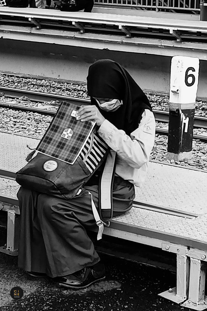 A student in school uniform sitting on a long concrete bench right next to the train tracks.