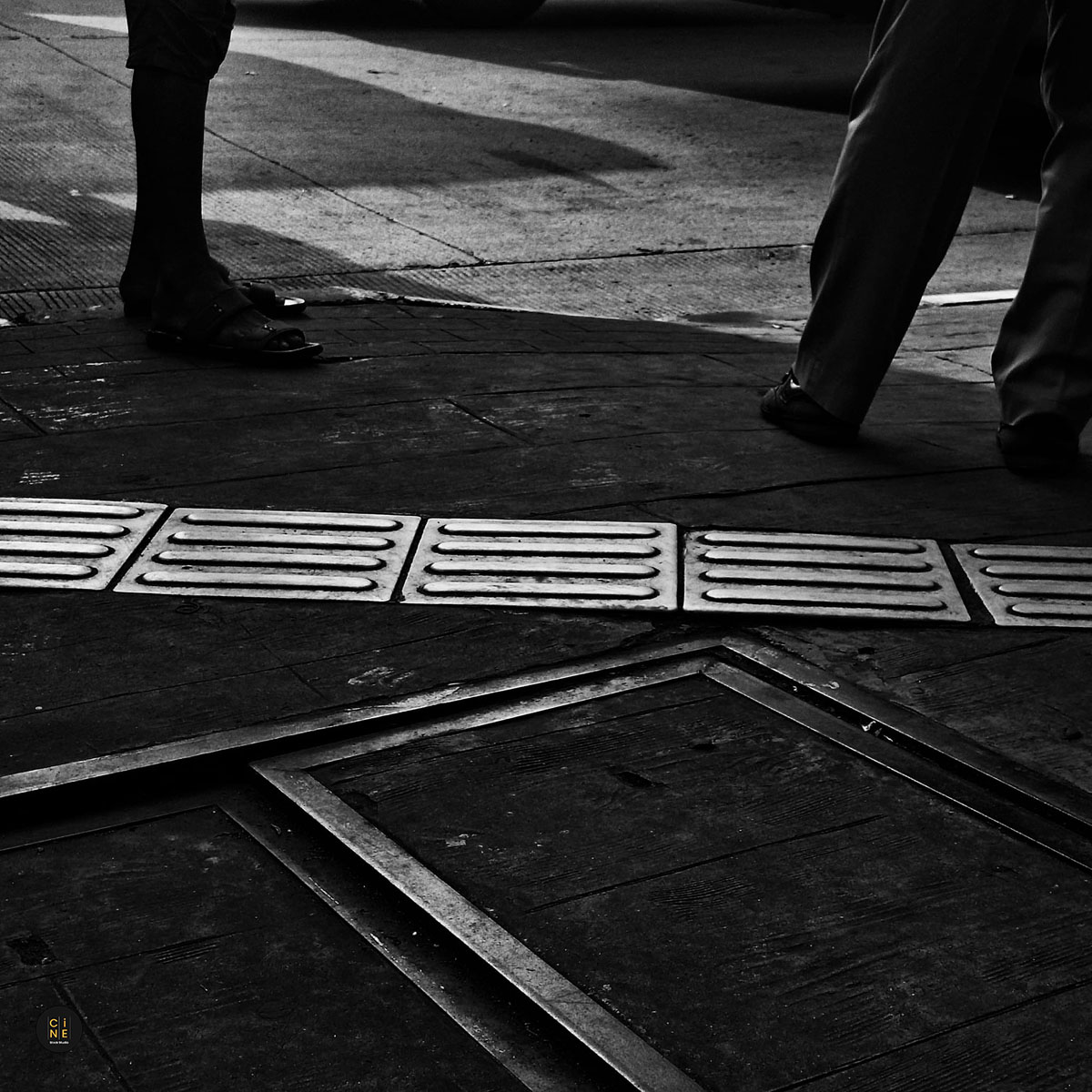 A high-angle monochrome shot of pedestrians walking across tactile paving guiding blocks on a concrete sidewalk.