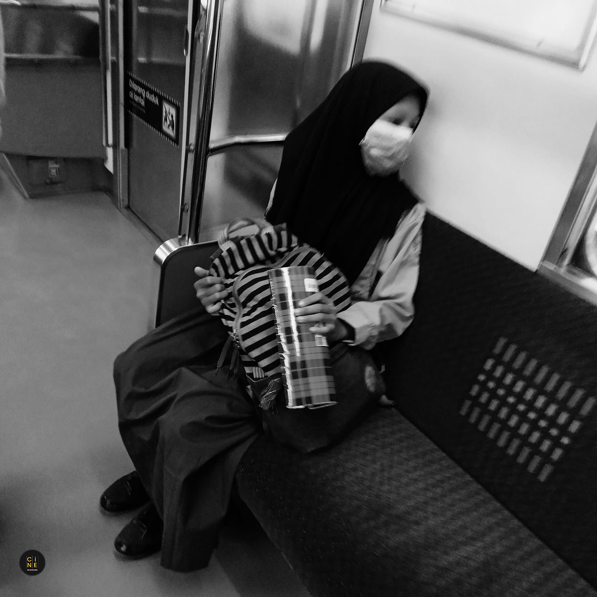 A student wearing a mask and hijab sitting alone on a train seat looking tired.