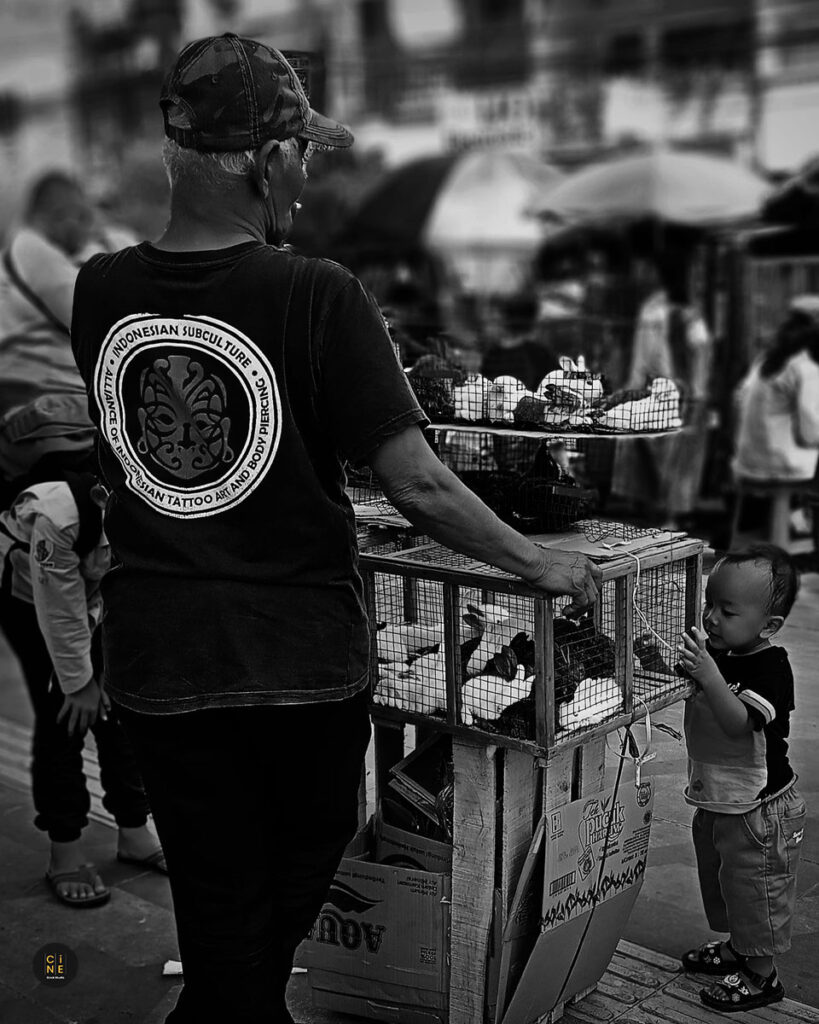 A wide-angle black and white street shot showing the rabbit vendor by his cart with a small child standing close to the cage.
