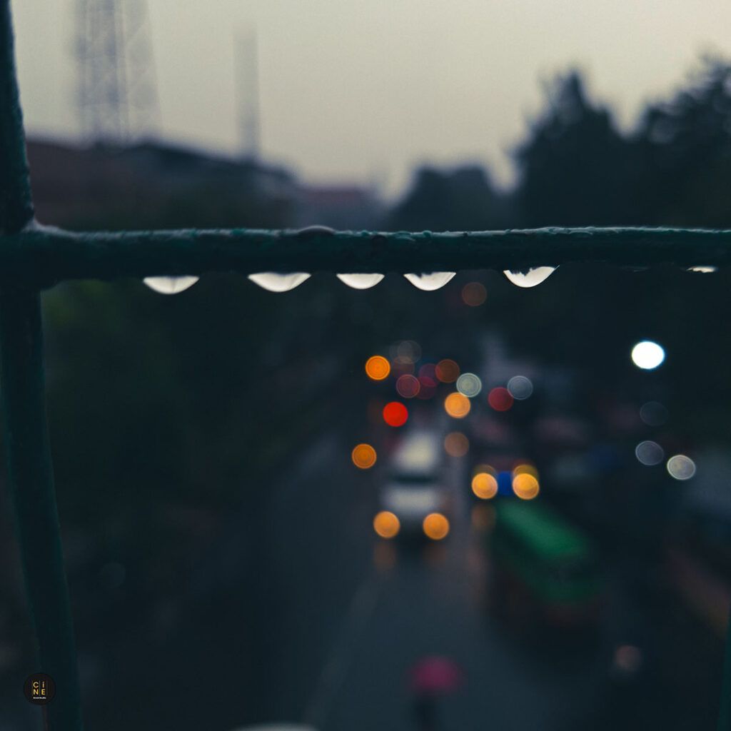 A vertical frame of crystal-clear water droplets clinging to an iron bar with vibrant city traffic bokeh lights in the background.