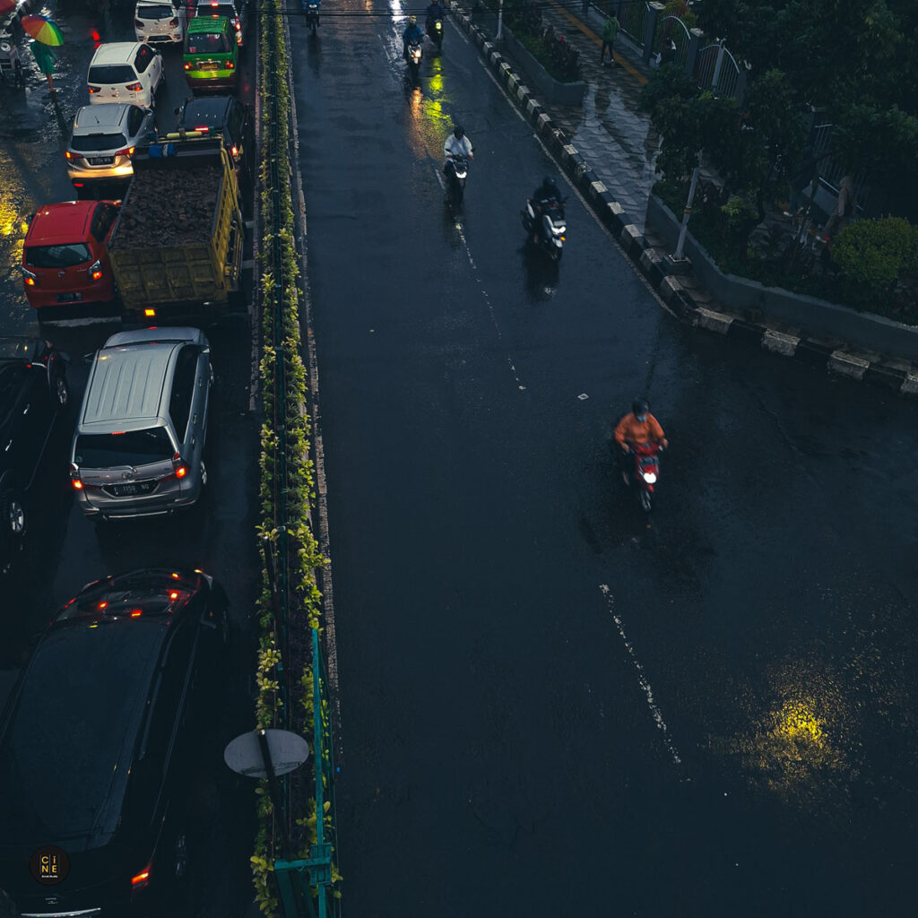 A high-angle street shot of a lone motorcycle riding on a dark, wet asphalt road reflecting headlights and street lamps.