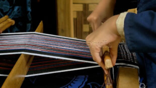 The technical side of weaving, focusing on the tools that pass through the vertical threads. Detail of a weaver's hand adjusting the bamboo shuttle on a loom