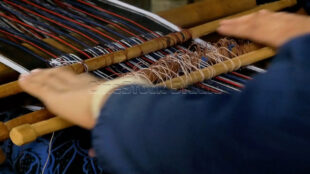 Capturing the rhythm and continuous movement of the weaving process in real-time. Close-up of hands moving across the loom with a soft motion blur